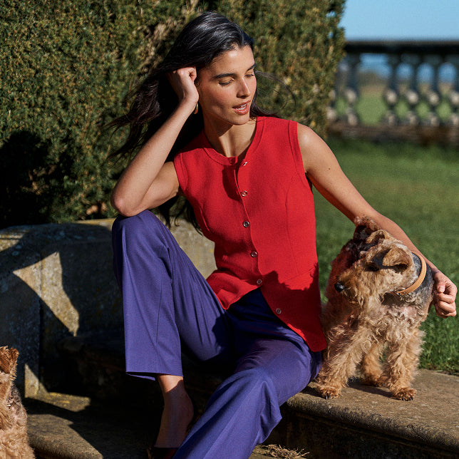 Eine Frau, die ein ärmelloses rotes Oberteil mit Knöpfen und eine lila Hose trägt, sitzt im Freien neben einem Hund, im Hintergrund sind Grünpflanzen und eine Steinmauer zu sehen.