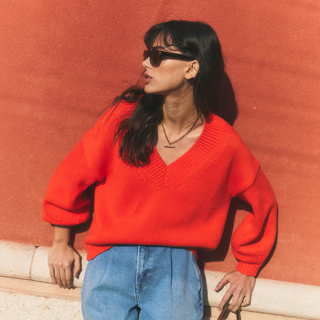 Eine Person, die einen knallroten Pullover und eine Sonnenbrille trägt, vor einer roten Wand.