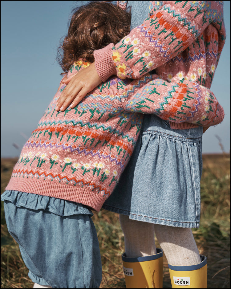 Girls hugging  in a field, wearing matching pink Boden Fair Isle cardigans