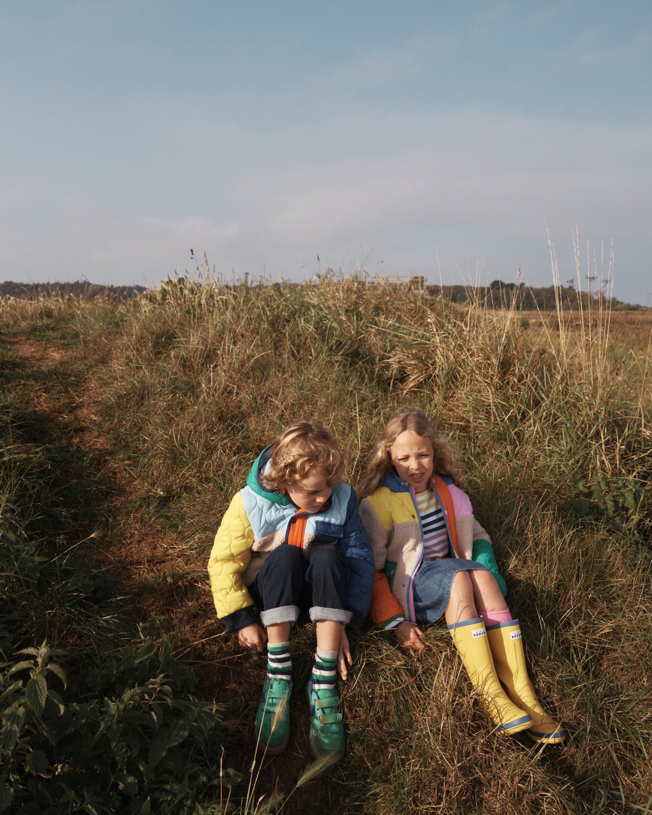 Girl and boy sat in a field, wearing colourful Boden borg coats