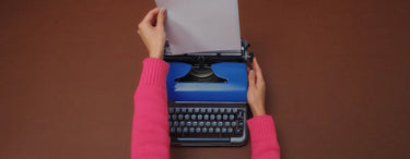 Eine Person, die auf einer blauen Schreibmaschine mit rosa Abdeckung vor einem braunen Hintergrund tippt