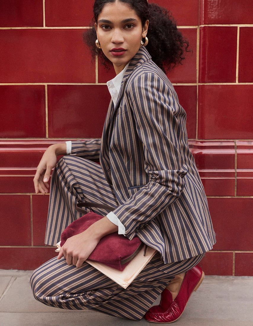 Ein Model hockt vor einer rot gefliesten Wand. Es trägt einen braun-beigen Nadelstreifenblazer und eine dazu passende Hose, hält eine burgunderrote Clutch und ein helles Buch in der Hand und trägt rote Leder-Loafer.