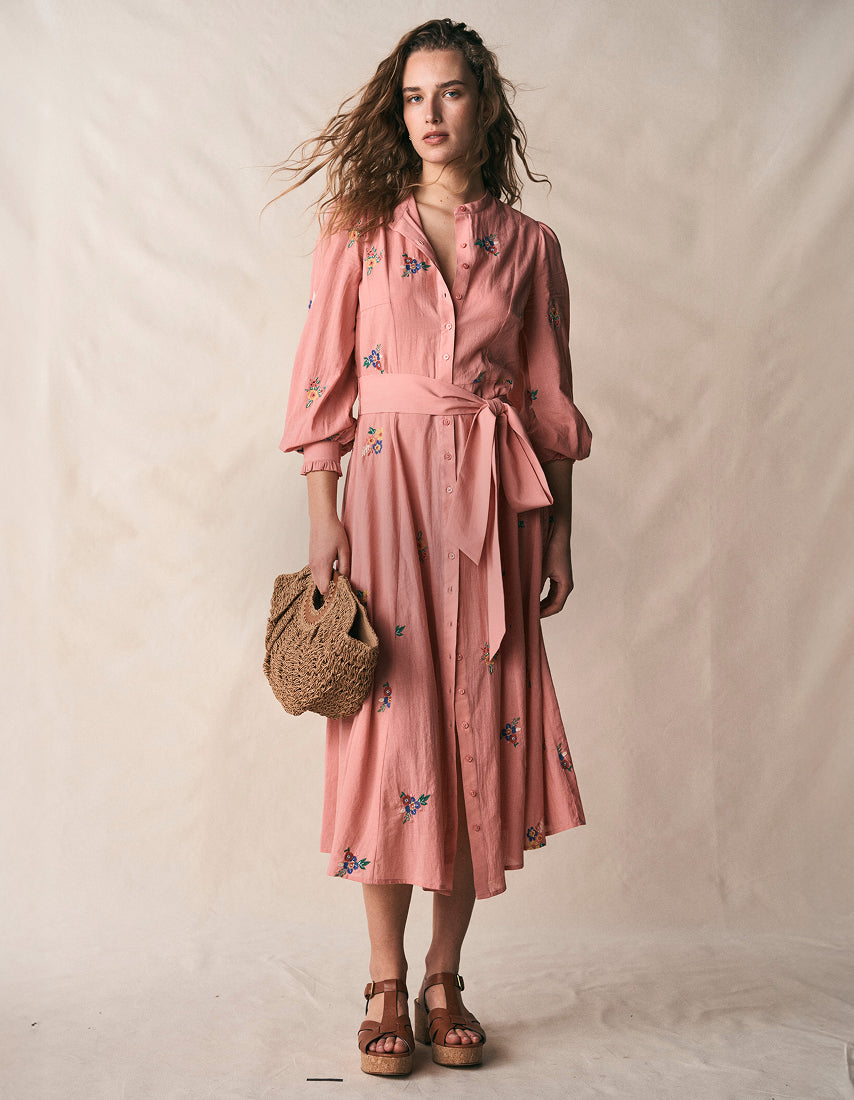 Eine Frau in einem rosa Kleid mit Blumenmuster, die eine geflochtene Tasche in der Hand hält, vor einem einfarbigen Hintergrund