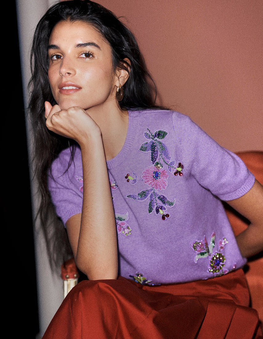 Eine Frau posiert in einem lavendelfarbenen, kurzärmeligen Strickpullover mit bunten Blumenstickereien und einem rostfarbenen Satinsrock vor einem warmen Terrakotta-Hintergrund.