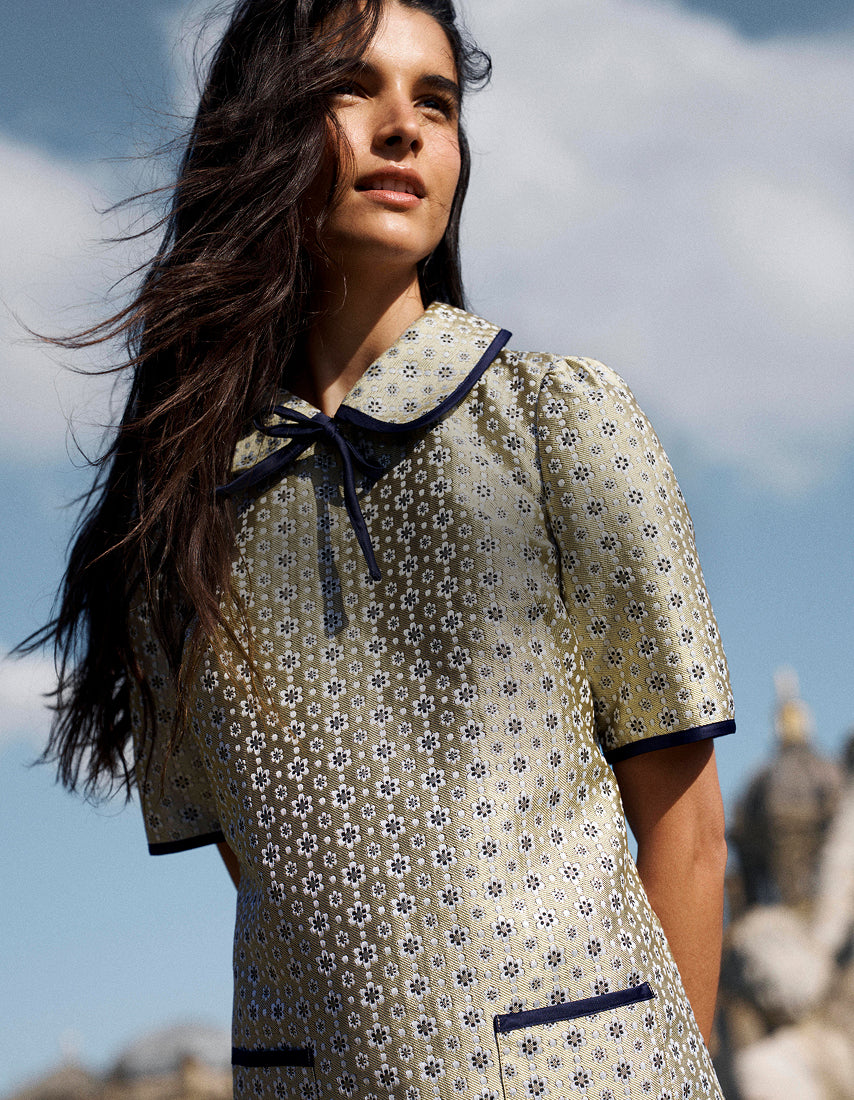 Eine Frau in einem cremefarbenen, kurzärmeligen Kleid mit einem kleinen blau-grünen Blumenmuster, einem dunkelblauen Kragen mit Krawatte und dunkelblauen Paspeln, fotografiert im Freien vor blauem Himmel.