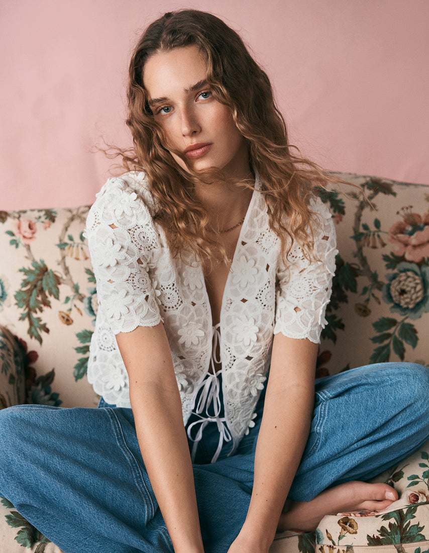 Eine Frau in einem weißen Spitzenoberteil und blauen Jeans sitzt auf einem blumigen Sofa.