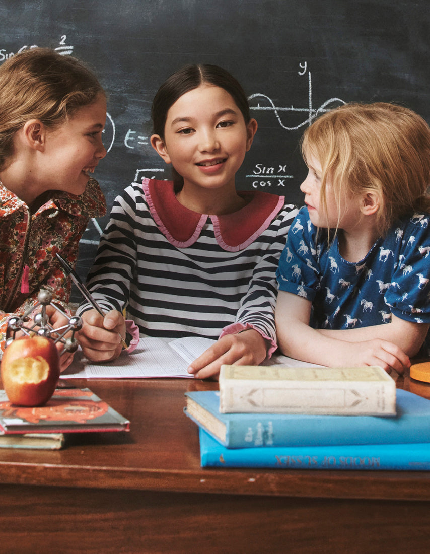 Drei Kinder sitzen an einem Schreibtisch mit Büchern und einem Apfel vor einer Tafel mit mathematischen Gleichungen.