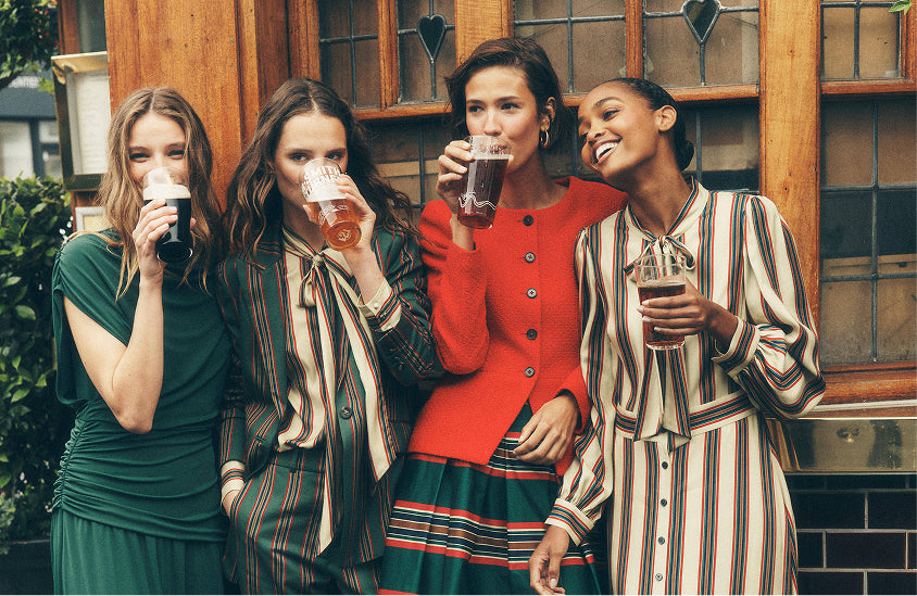 Vier Frauen stehen gemeinsam im Freien vor einem Holzgebäude und halten jeweils ein Pint Bier in der Hand. Sie tragen farbenfrohe, gemusterte Outfits, darunter ein dunkelgrünes Kleid, gestreifte Blusen und eine leuchtend rote Strickjacke.