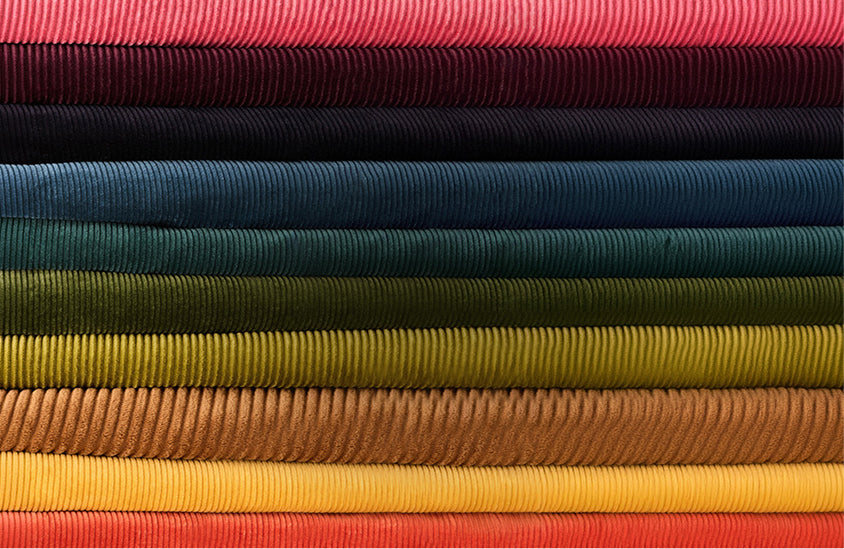 Eine Nahaufnahme eines Stapels gerippter Stoff- oder Materialmuster in verschiedenen Farben, darunter Rosa, Marineblau, Blaugrün, Grün, Olivgrün, Hellbraun, Senfgelb, Gelb, Orange und Rot, die horizontal angeordnet sind.