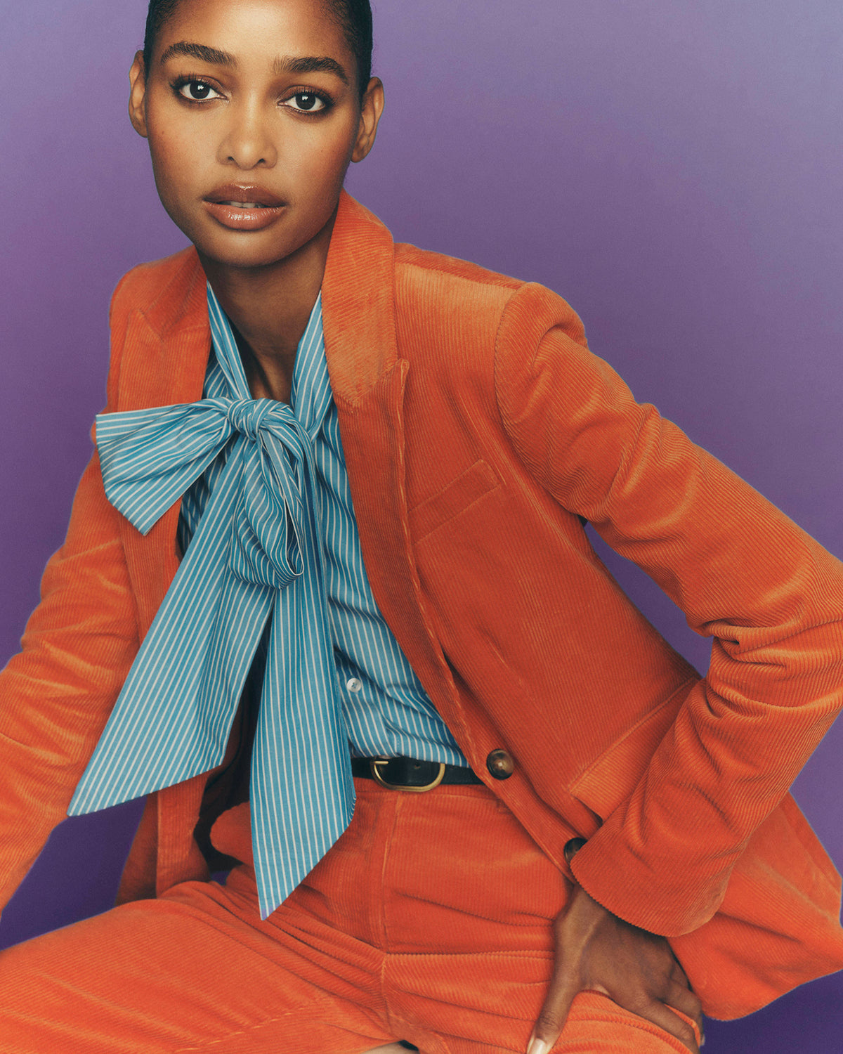 Ein Model trägt einen leuchtend orangefarbenen Cordblazer und eine dazu passende Hose, dazu ein hellblau-weiß gestreiftes Hemd mit einer großen Fliege, vor einem einfarbigen violetten Hintergrund.