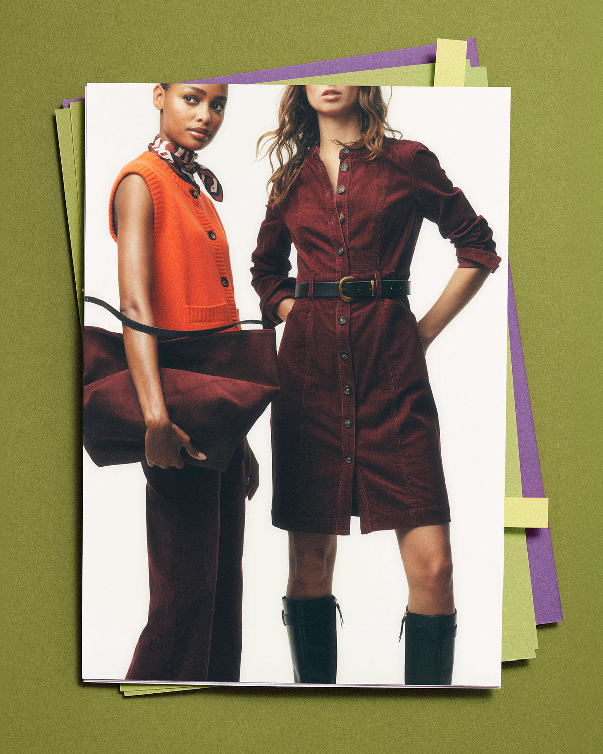 Eine Seite aus einem Modemagazin oder -katalog, auf der zwei Models zu sehen sind: Das eine trägt eine orangefarbene Weste und eine braune Hose und hält eine braune Tasche in der Hand, das andere ein dunkelrotes Cord-Hemdkleid mit einem schwarzen Gürtel und schwarzen Stiefeln. Die Seite ist auf mehreren bunten Papierbögen vor einem einfarbigen olivgrünen Hintergrund abgebildet.
