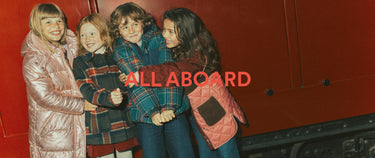 Vier Kinder, die sich vor einem roten Zug umarmen und bunte Winterjacken tragen. Über dem Bild ist in roter Schrift der Schriftzug „ALL ABOARD“ zu lesen.