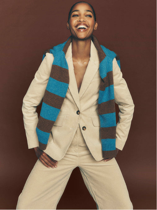 Ein lächelndes Model, das einen cremefarbenen Blazer und eine dazu passende Hose trug, kombiniert mit einer blau-braun gestreiften Strickweste, posierte vor einem einfarbigen dunkelbraunen Hintergrund.