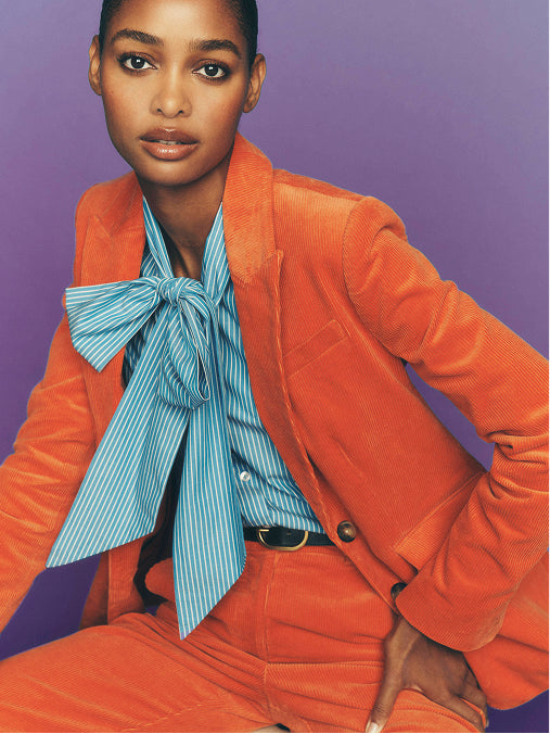 Ein Model trägt einen leuchtend orangefarbenen Cordblazer und eine dazu passende Hose, dazu ein hellblau-weiß gestreiftes Hemd mit einer großen Fliege, vor einem einfarbigen violetten Hintergrund.