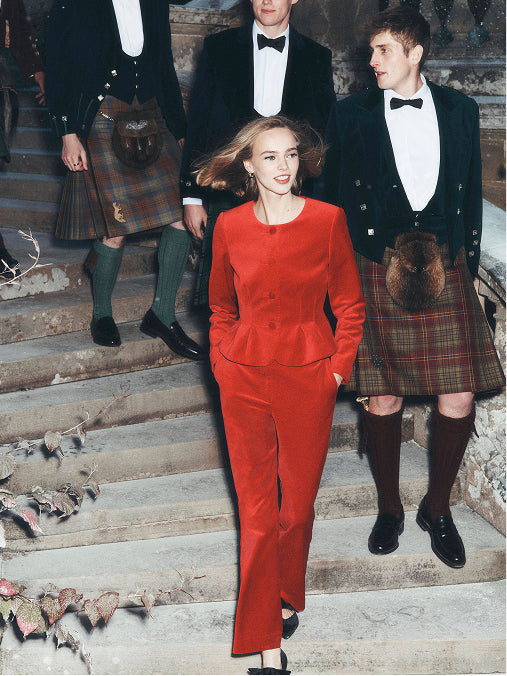 Eine Frau in einem knallroten, maßgeschneiderten Kostüm geht eine Steintreppe hinunter, während drei Männer in schwarzen Smokings und Tartan-Kilts hinter ihr stehen.