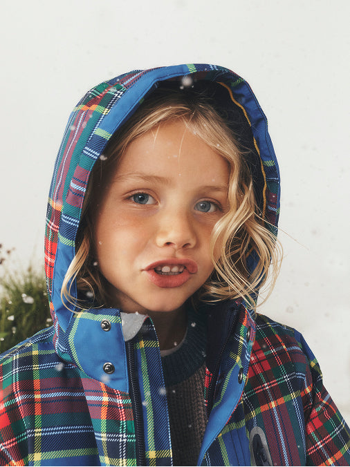 Ein kleines Kind, das eine karierte Kapuzenjacke mit blauem Besatz trägt, fotografiert im Freien vor einem hellen, neutralen Hintergrund, während Schneeflocken fallen.