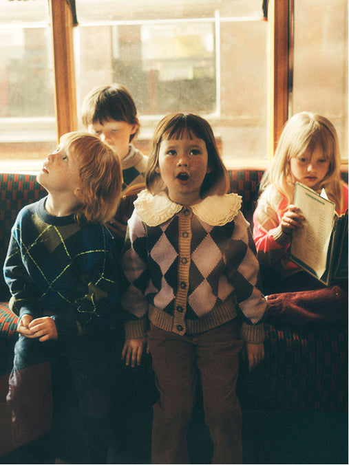 Ein Foto im Vintage-Stil, das vier Kinder zeigt, die in einem Zug oder Bus sitzen. Das Kind in der Mitte trägt einen braun-beigen Argyle-Pullover mit weißem Spitzenkragen, während das Kind links einen dunkelblauen Argyle-Pullover trägt und das Kind rechts ein Buch in der Hand hält.
