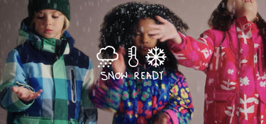 Drei Kinder in bunten Jacken und Hosen stehen im Schnee und lächeln, gekleidet in Boden Kleidung.