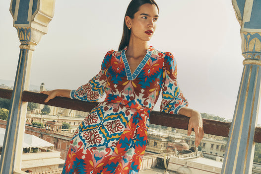Eine Frau in einem bunten Kleid steht auf einem Balkon, im Hintergrund ist eine Stadtlandschaft zu sehen