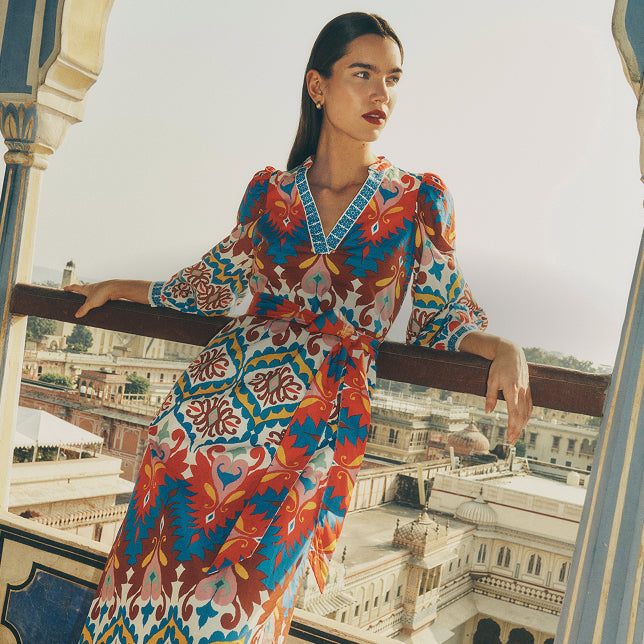 Eine Frau in einem bunten Kleid steht auf einem Balkon, im Hintergrund ist eine Stadtlandschaft zu sehen