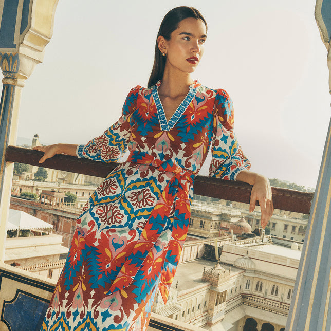 Eine Frau in einem bunten Kleid steht auf einem Balkon, im Hintergrund ist eine Stadtlandschaft zu sehen