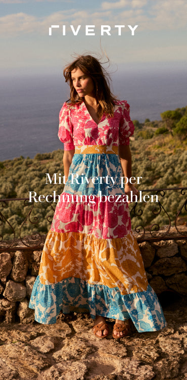 Mit Riverty per Rechnung bezahlen: ERFAHREN SIE MEHR - Woman in a coluorful floral dress standing by a stone railing with a sea view.