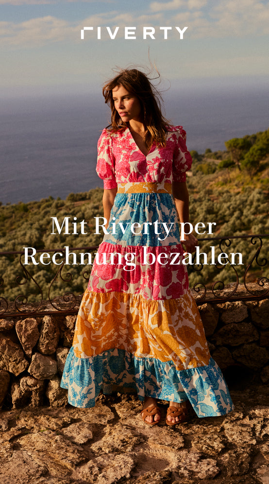 Mit Riverty per Rechnung bezahlen: ERFAHREN SIE MEHR - Woman in a coluorful floral dress standing by a stone railing with a sea view.