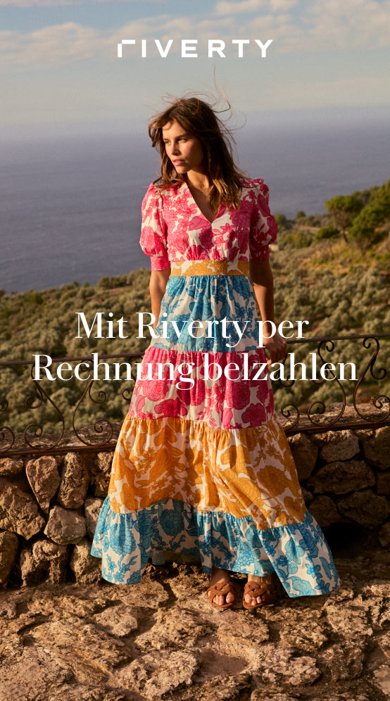 Mit Riverty per Rechnung bezahlen: ERFAHREN SIE MEHR - Woman in a coluorful floral dress standing by a stone railing with a sea view.